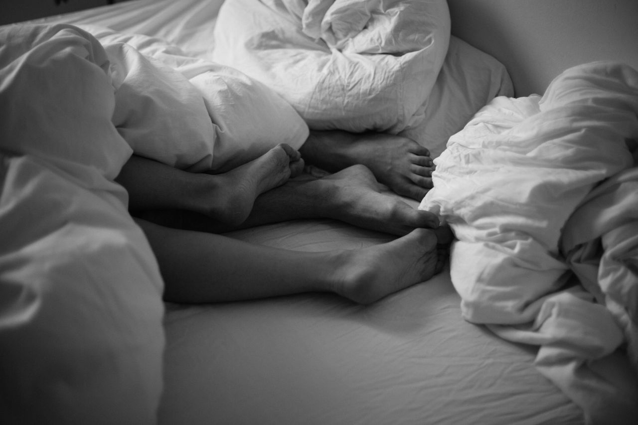 our-story-01 Black and white image of feet intertwined under cozy bedsheets, capturing a peaceful moment.