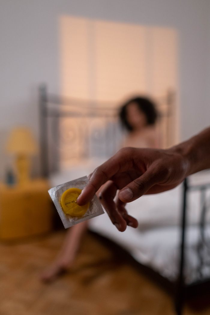 A hand holding a condom in focus with a blurred background of a person sitting on a bed.